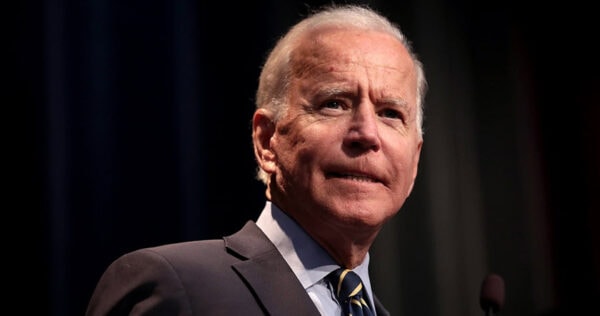 An older man in a suit and tie stands at a podium, looking forward with a neutral expression, against a dark background.