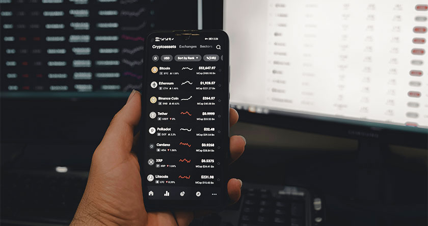 A person holds a smartphone displaying cryptocurrency prices in front of two computer monitors showing data charts and tables.