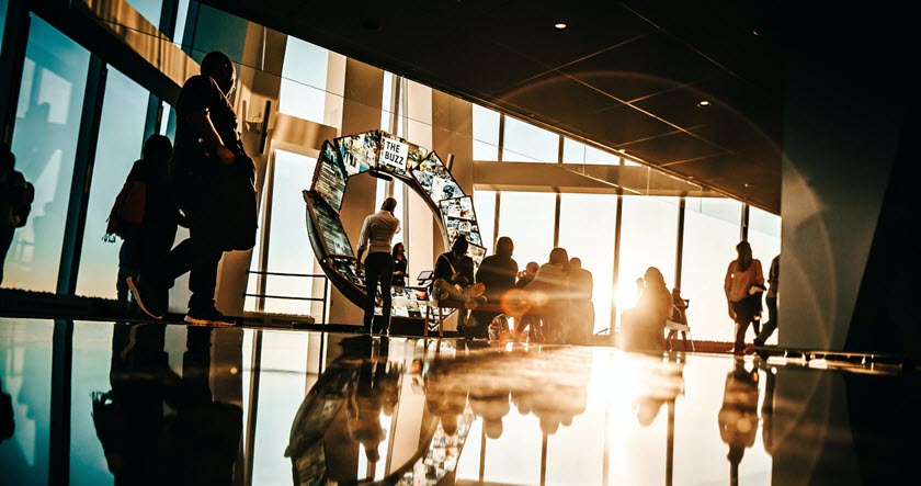 People are gathered near a large window with sunlight streaming in, some sitting and some standing, with a curved digital display in the background.