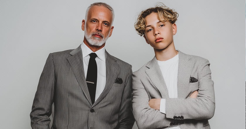 Two men wearing suits stand side by side against a plain background; the older man has gray hair, and the younger man has curly blond hair and folded arms.